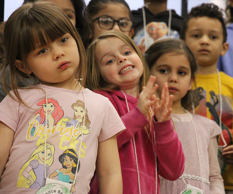 A group of kindergarten students standing together singing. The girl in the center is smiling broadly and wearing a dark pink shirt.