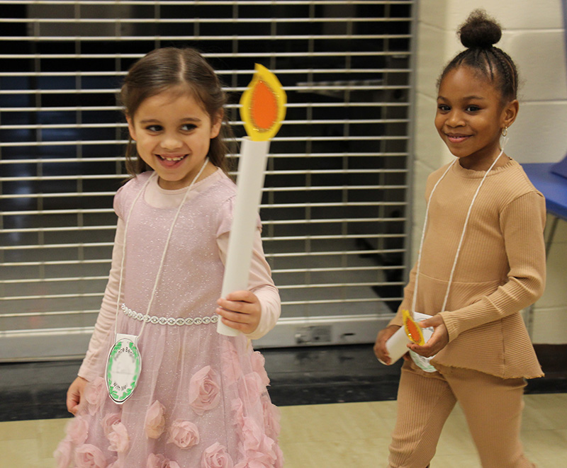 Two kindergarten girls walk into a room smiling. Each is holding a big paper candle.