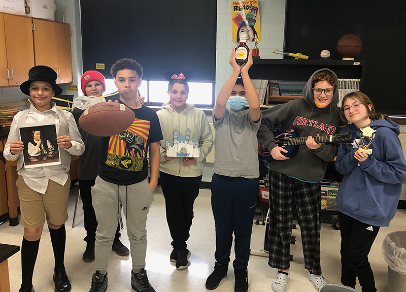 A group of seven middle school students stand in a line holding props representing the person they studied. one student has a top hat on and is holding a picture of his subject, another is holding a football, one is holding a cut-out of a Disney castle, one has a jar of barbeque sauce over head, one is holding a guitar and another has a trophy.