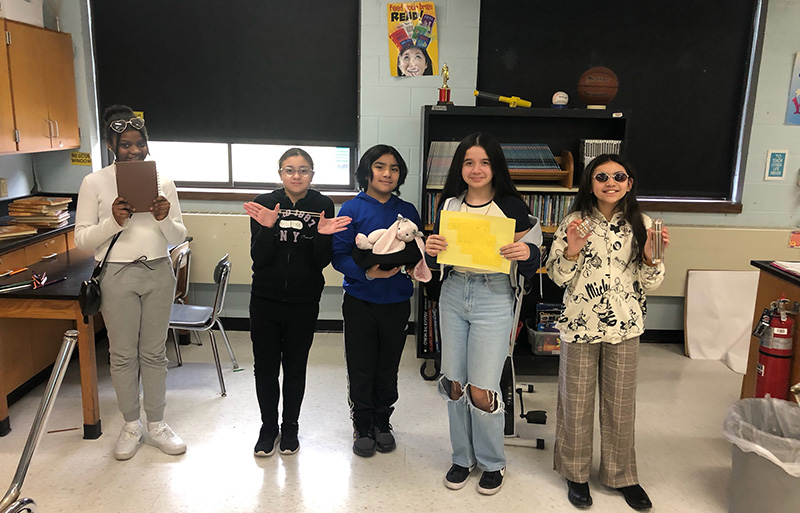 Five middle school students stand together either dressed as the person they studied or holding items representing that person. From left a student is holding a diary, an elastic, a bunny,  a yellow piece of paper and the last is dressed in funky clothes, round sunglasses.