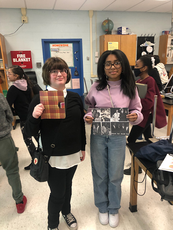 Two sixth-grade girls, both with glasses, stand together. One is holding a diary and the other pictures of the singer Aretha Franklin.