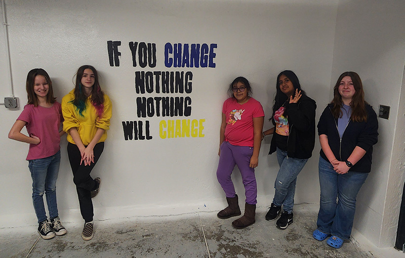 Five middle school students stand in front of a white wall on which they painted "If you Change Nothing, Nothing will change" in black, blue and yellow.