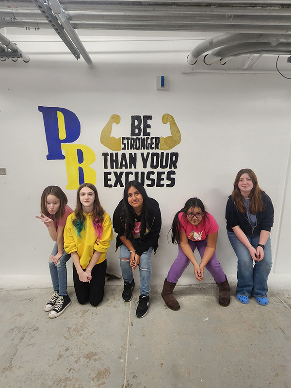 Five middle school students squat down in front of a mural they painted on a white wall. It says PB Be stronger than your excuses. Arms flexing are coming out of the word stronger.