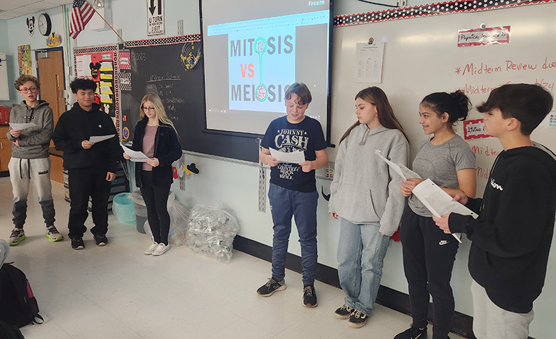 Two groups of students - three on the left and four on the right - stand in front of a classroom to debate. In the center is a slide that says Mitosis vs. Meiosis.