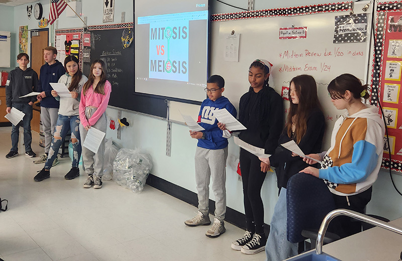 Two groups of students - four on the left and four on the right - stand in front of a classroom to debate. In the center is a slide that says Mitosis vs. Meiosis.
