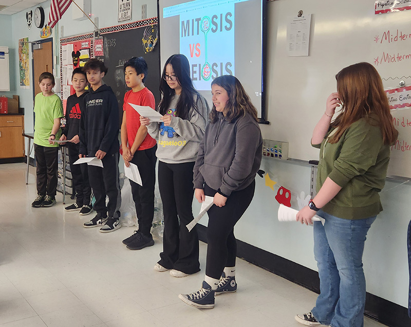 Two groups of students - four on the left and three on the right - stand in front of a classroom to debate. In the center is a slide that says Mitosis vs. Meiosis. One student near the center is reading from a piece of paper.