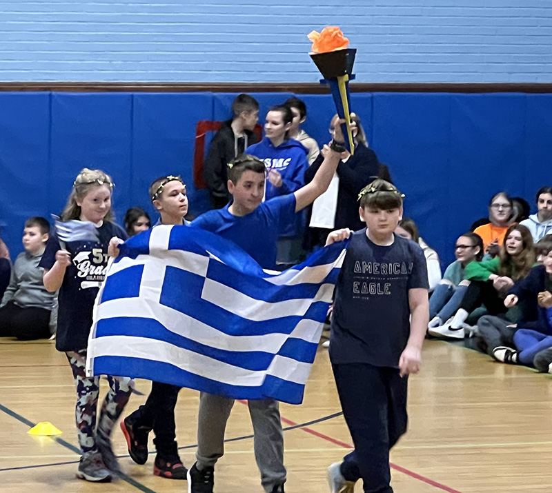 Three middle school students carry the blue and white flag of Greece into the gym. another student holds up a paper torch.