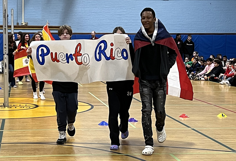 Three middle school students parade in with a sign that says Puerto rico. One student is wearing the Puerto Rican flag as a cape.