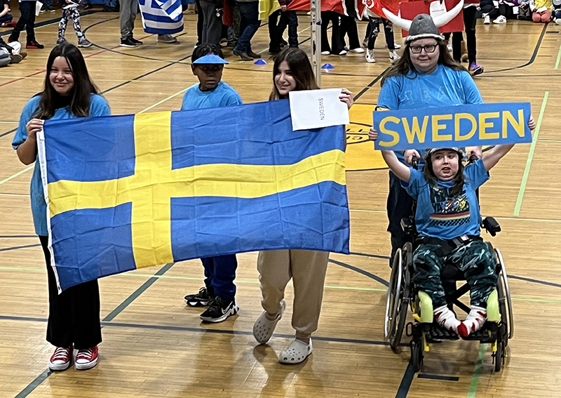 Three middle school students carry the flag of Sweden, while another student holds a sign saying Sweden over her head. She is in a wheel chair being pushed by a student.