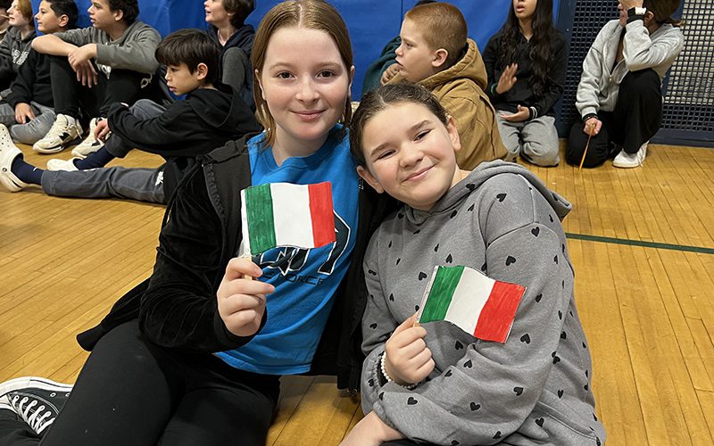 A teacher serves during the volleyball game with students watching in background