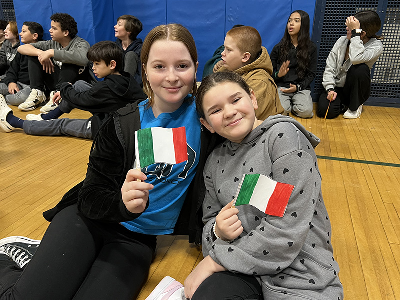 Two middle school girls sit together on a gym floor and hold up small flags of Italy.