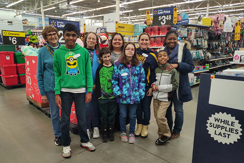 A group of four middle school students stand in front of five female adults. They are standing in front of lots of retail merchandise 