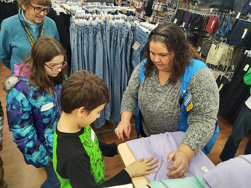 A woman with shoulder-length dark hair, wearing a black and white shirt and blue vest, helps two students who are using a board to fold a shirt.