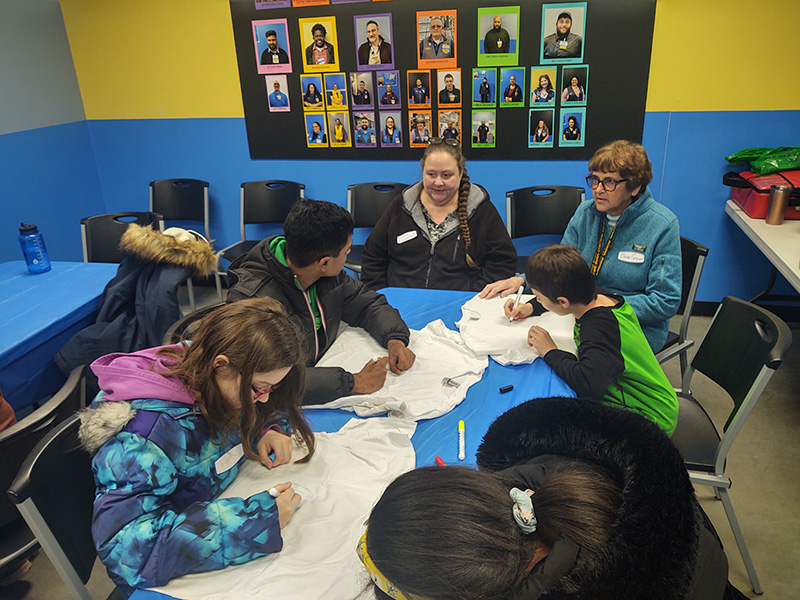 A group of four middle school students sit at a table with two adults decorating tshirts.