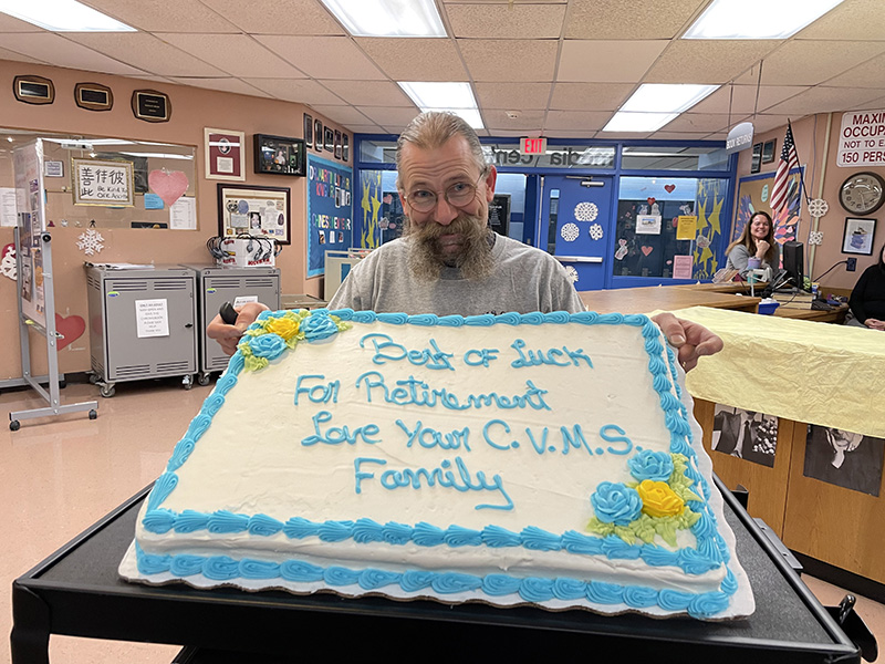 A man wearing a gray shirt with his long hair pulled back in a pony tail and a large handlebar mustache holds a big sheet cake. The cake is white with blue edging and writing.The cake says Good Luck in Retirement Love your CVMS Family.