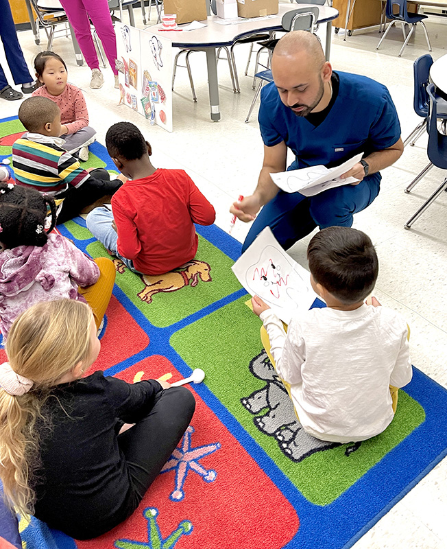Younger elementary students sit on a colorful rug with worksheets containing a picture of a large tooth on it. A man in dark blue scrubs kneels down to talk to one of the students about his sheet.