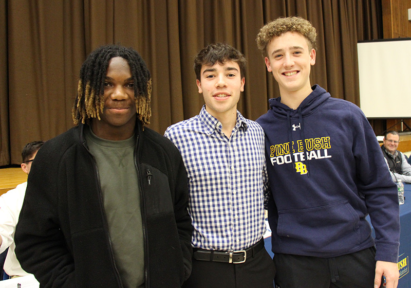 Three high school age young men stand together smiling. There is a curtain and stage behind them.
