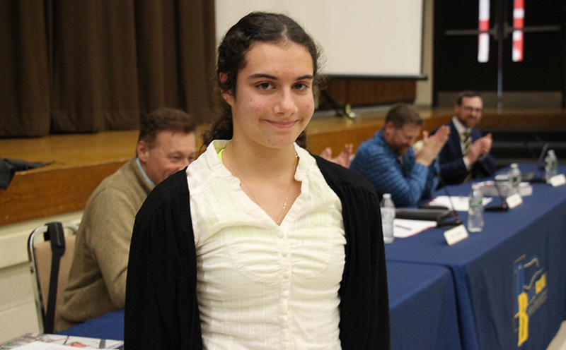 A high school age young woman, whose long dark hair is pulled back into a ponytail. Stands smiling with people behind her clapping. She is wearing a white blouse and black sweater.