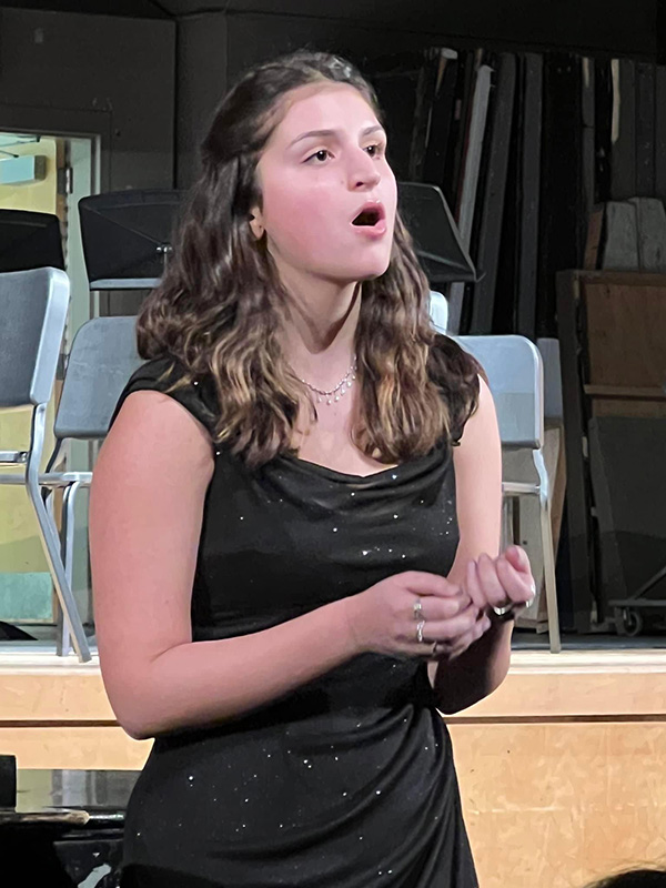 A high school senior girl with long brown hair, wearing a black sparkly dress sings.