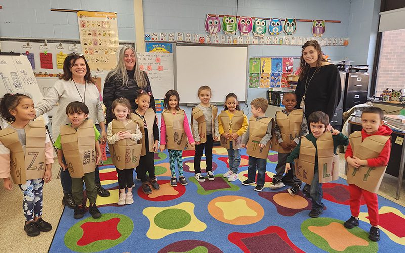 Eleven kindergarten students stand in front of three adult women. The kids have brown paper vests on, each with letters on them. They are standing on a rug with multi colors.