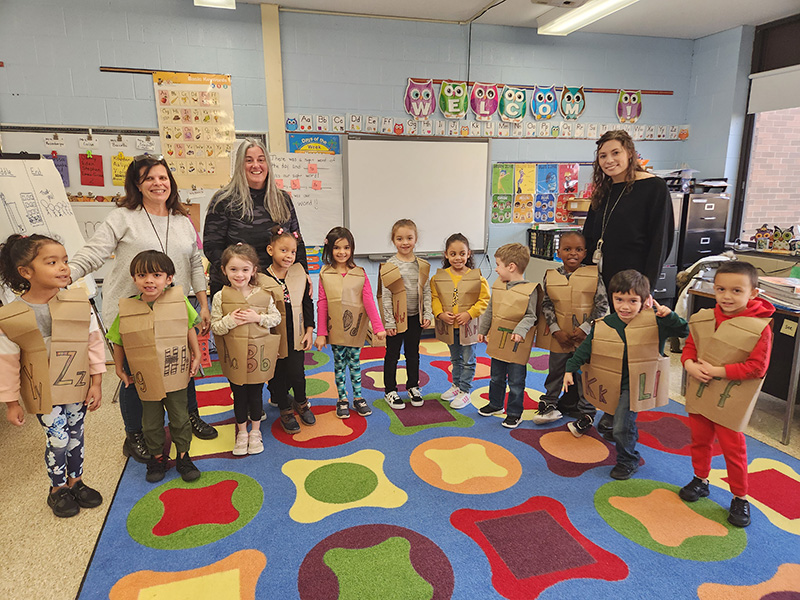Eleven kindergarten students stand in front of three adult women. The kids have brown paper vests on, each with letters on them. They are standing on a rug with multi colors.