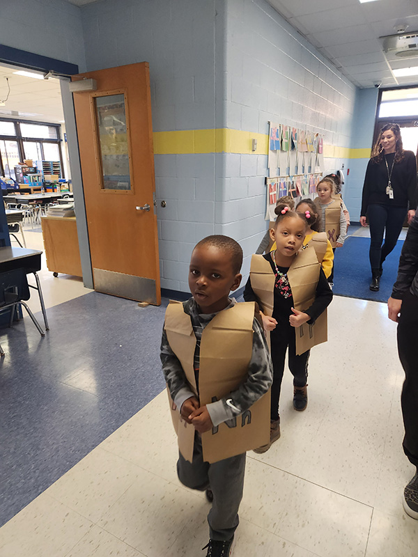 kindergarten students march through the hallway. They are wearing brown paper vests with letters on them.