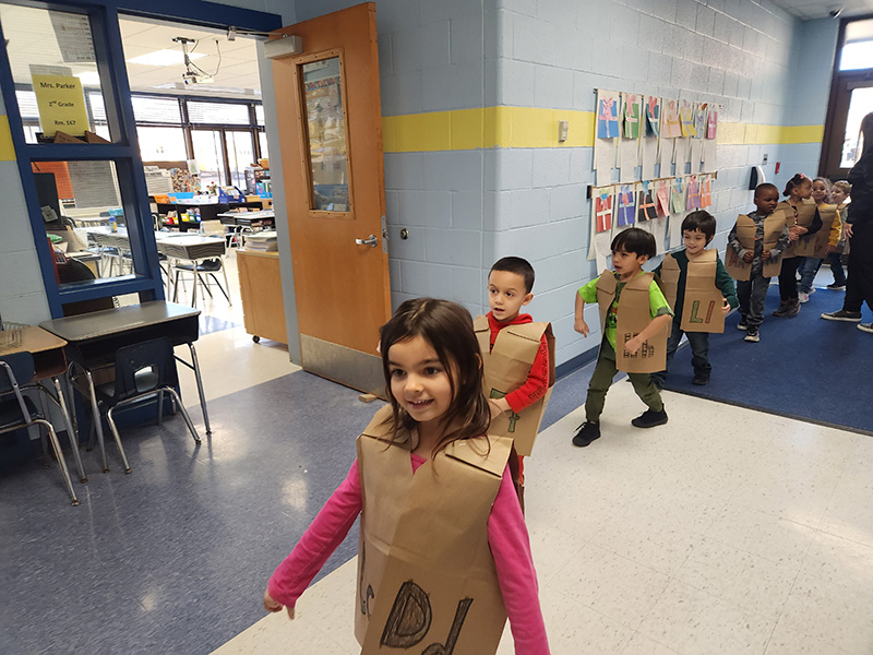 kindergarten students march through the hallway. They are wearing brown paper vests with letters on them.