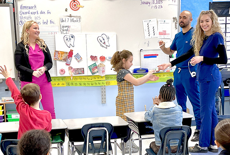A woman with long blonde hair, wearing pink scrubs and a black sweater, stands in front of a class on the left. A young elementary student stands in the center, and two other adults, both dressed in blue scrubs, stand on the right. Two other students sit at desks, one has his hand up.