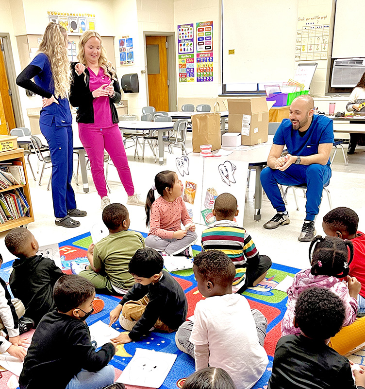 A group of younger elementary students sit on a colorful rug and listen to three adults, all wearing scrubs, talking about dental health.
