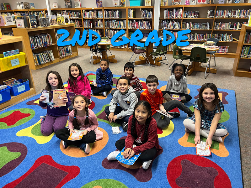 A group of 10 second-grade students sit on a colorful rug. Each is holding a book.