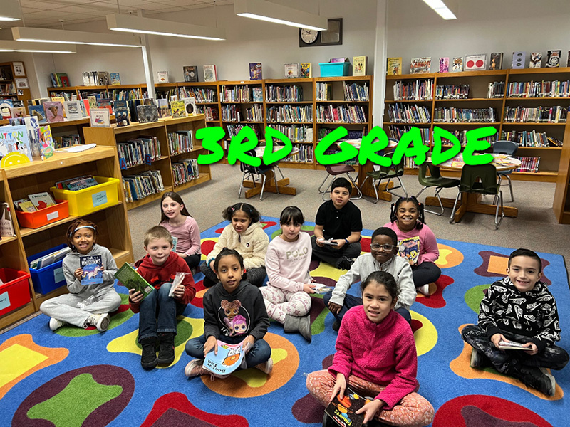 A group of 11 third-grade students sit on a colorful rug. Each is holding a book.