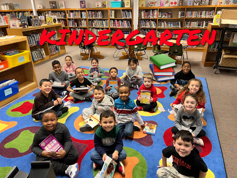 A group of 16 kindergarten children sit on a colorful rug. They are all holding books.
