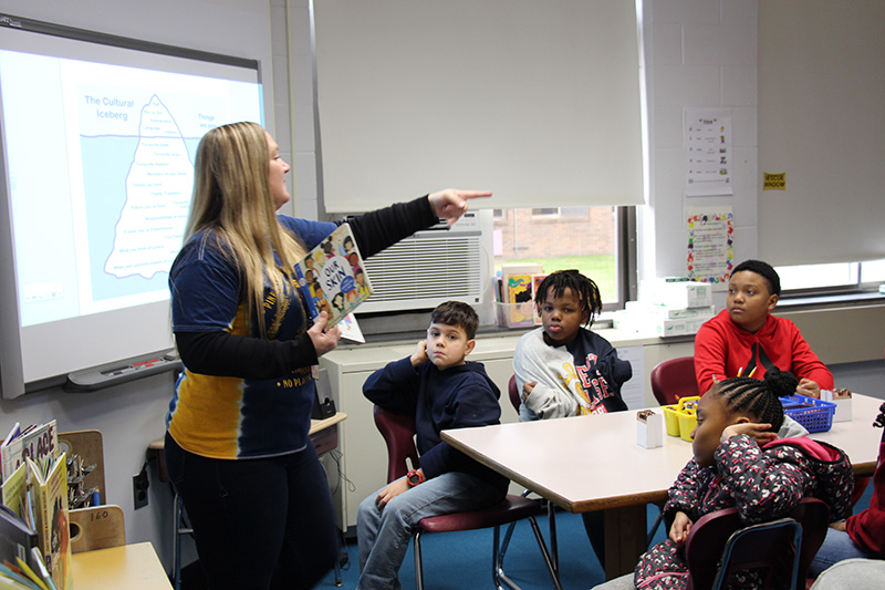 A teacher with long blonde hair, wearing a blue and gold shirt, points to a student to answer a questions. There are tables in front of her with fourth-grade students at them.