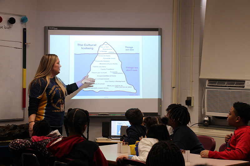 A teacher with long blonde hair stands at a projected image on a screen, which is an iceberg showing what is seen above the water and what's under the water.