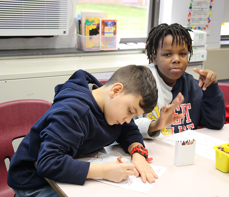 Two fourth-grade boys work at a table, coloring and writing on paper.