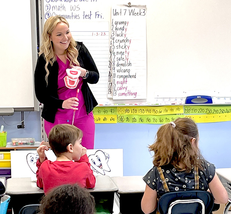 A woman with long blonde hair, wearing pink scrubs and a black sweater, stands in front of the class with a large plastic tooth and a large toothbrush, showing the students sitting in front of her how to brush correctly.