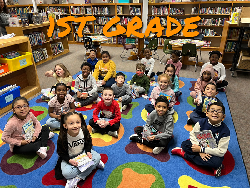 A group of 19 first-grade students sit on a colorful rug. Each is holding a book.