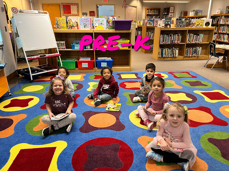 A group of six pre-K students sit smiling on a colorful rug.
