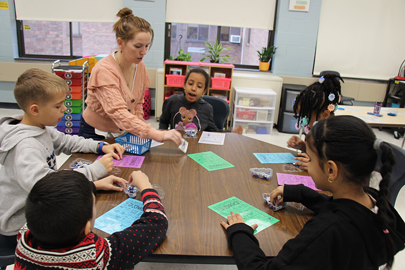 A woman wearing a peach colored sweater, her hair pulled up, sits at a table with five elementary students. They are playing bingo with pictures of winter items.