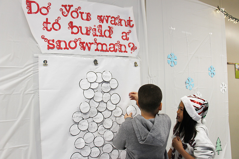Two elementary students stand at a wall that is covered in white fabric. In red letters at the top it says Do you want to build a snowman. One of the students in front of it is putting a white paper snowball on the tree.