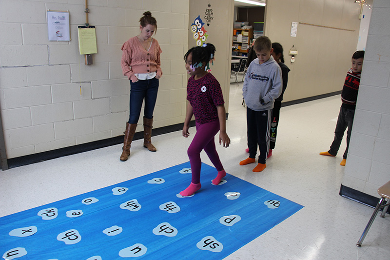 A blue mat is on the floor with white patches that have letters on them. A woman stands next to it as an elementary age girl has her feet on two different white patches. There are two other students behind her waiting for their turn.