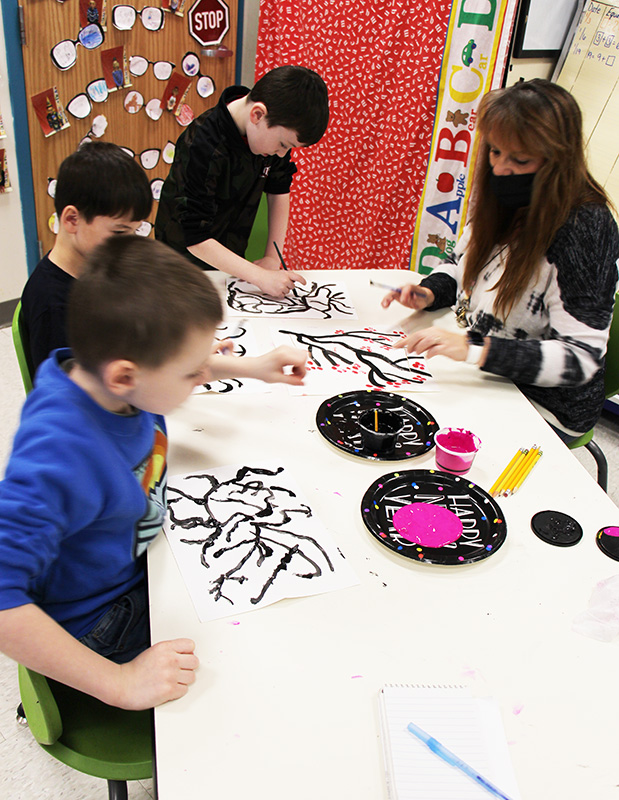 Three first-grade boys work at a table with a woman. They are making paintings of cherry blossoms.