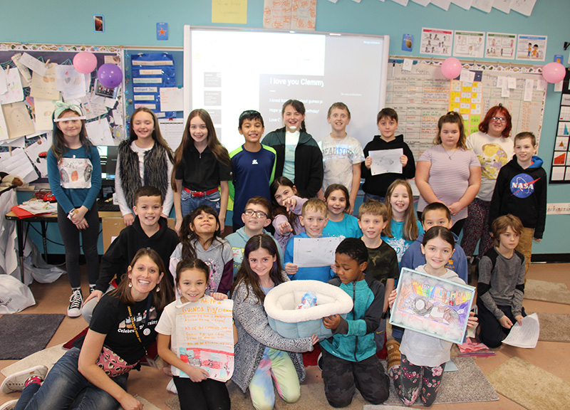 A group of 24 fourth-grade students standing and kneeling in three rows. In the front, two students are holding a cozy carrier with a guinea pig in it. There is one adult sitting on the floor. All are smiling.