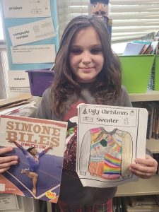 An elementary age girl with longer dark hair smiles as she holds up a book about Simone Biles and a page she colored.