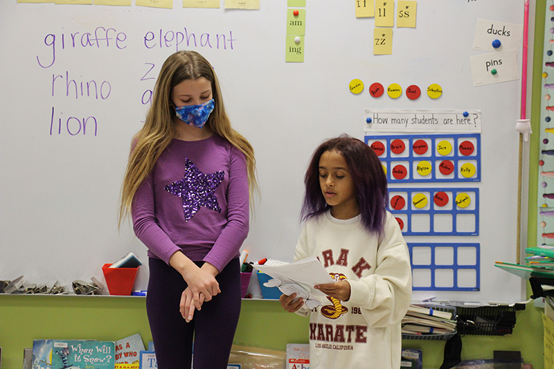 Two older elementary students stand in front of a class making a presentation. The student on the left is tall and has long blonde hair and is wearing dark pants and a light purple shirt and mask. The student on the right is shorter, with shoulder length dark hair and a white sweatshirt. 