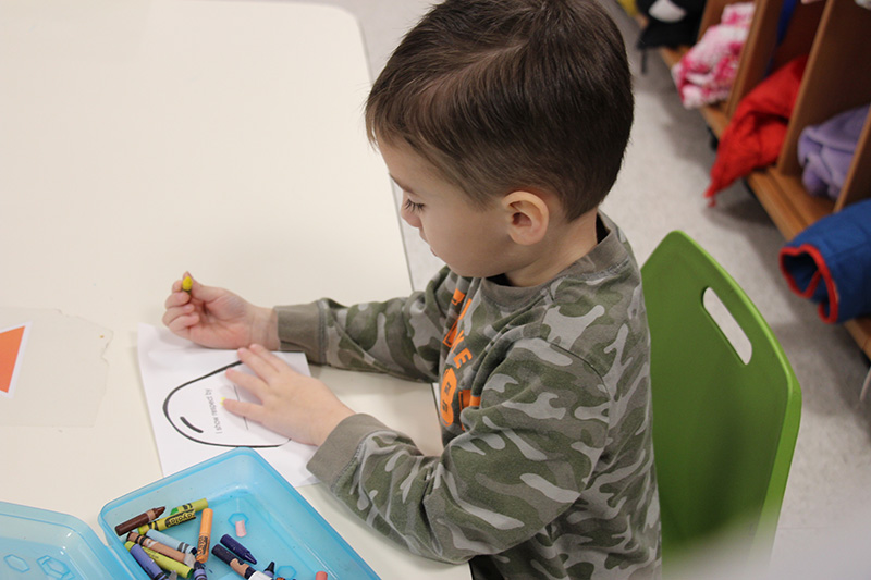 A pre-school boy with short dark hair wearing a green camouflage shirt  sits at a table coloring a paper gumdrop.