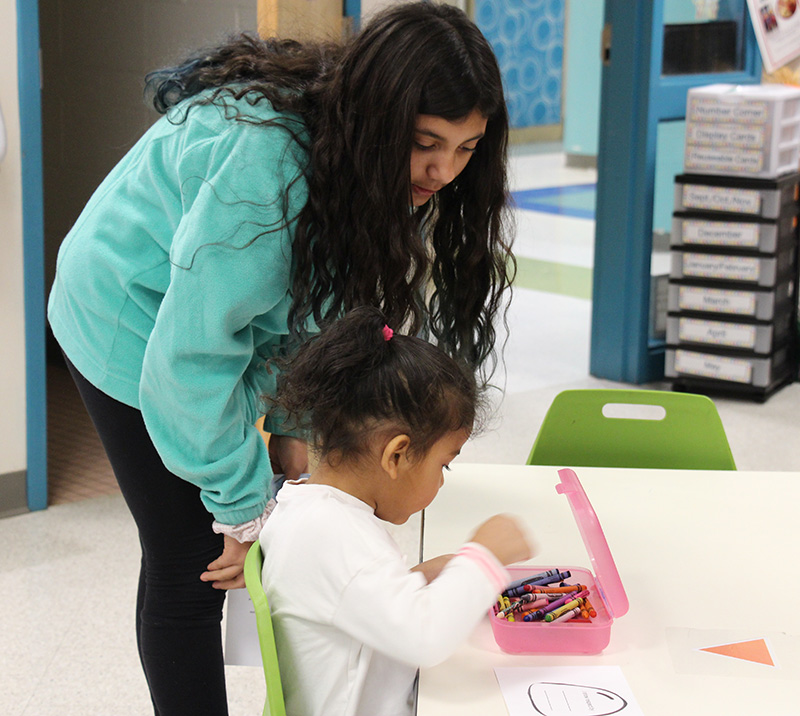 An older elementary student with long dark hair and light teal sweatshirt looks over a younger elementary student with long dark hair pulled into a ponytail who is coloring a paper gumdrop. 