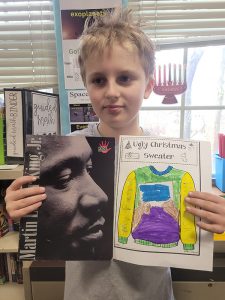 An elementary-age boy holds up a book about Martin Luther King Jr and a page he colored abouthim.