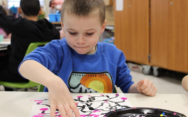 A first-grade boy, with a very short haircut, wearing a long-sleeve blue shirt, uses his thumb to put pink blossoms on a painting of twigs.