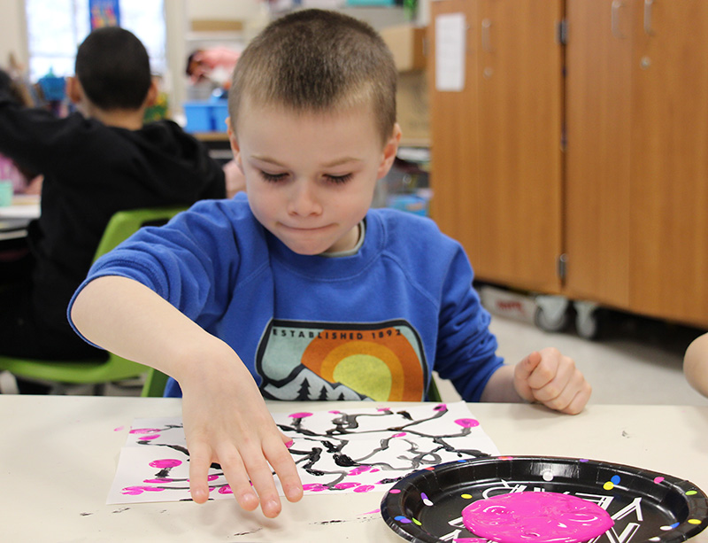 A first-grade boy, with a very short haircut, wearing a long-sleeve blue shirt, uses his thumb to put pink blossoms on a painting of twigs.